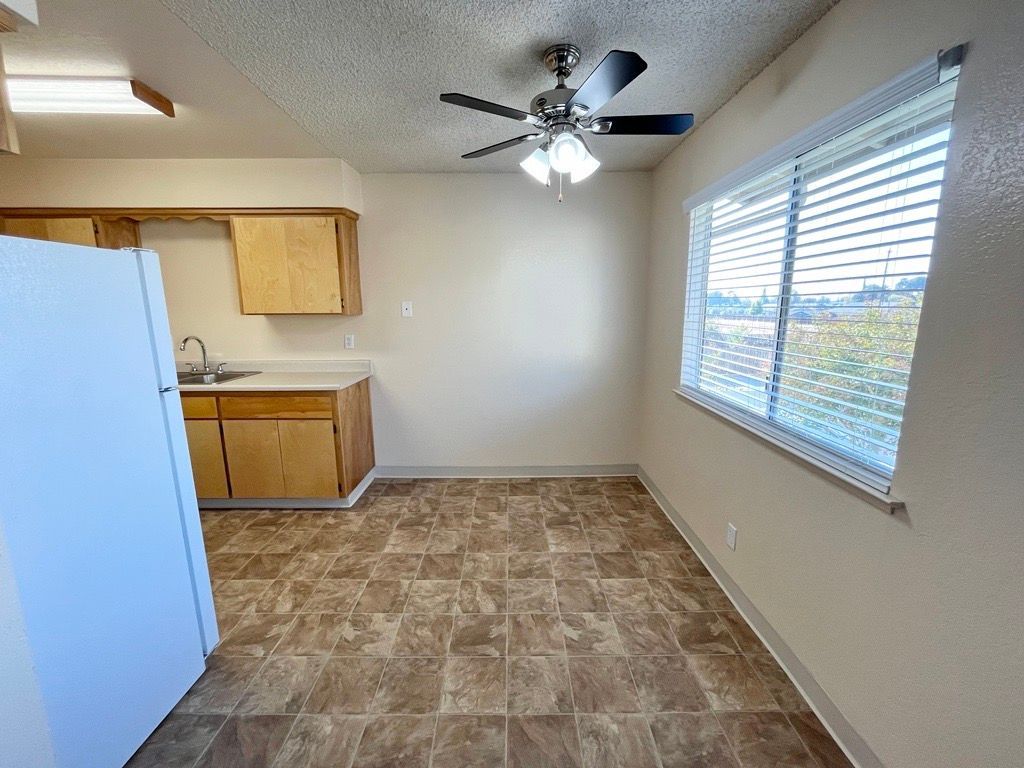 Kitchen area with cabinets, a refrigerator, and a window with blinds. Brown flooring and a ceiling fan.