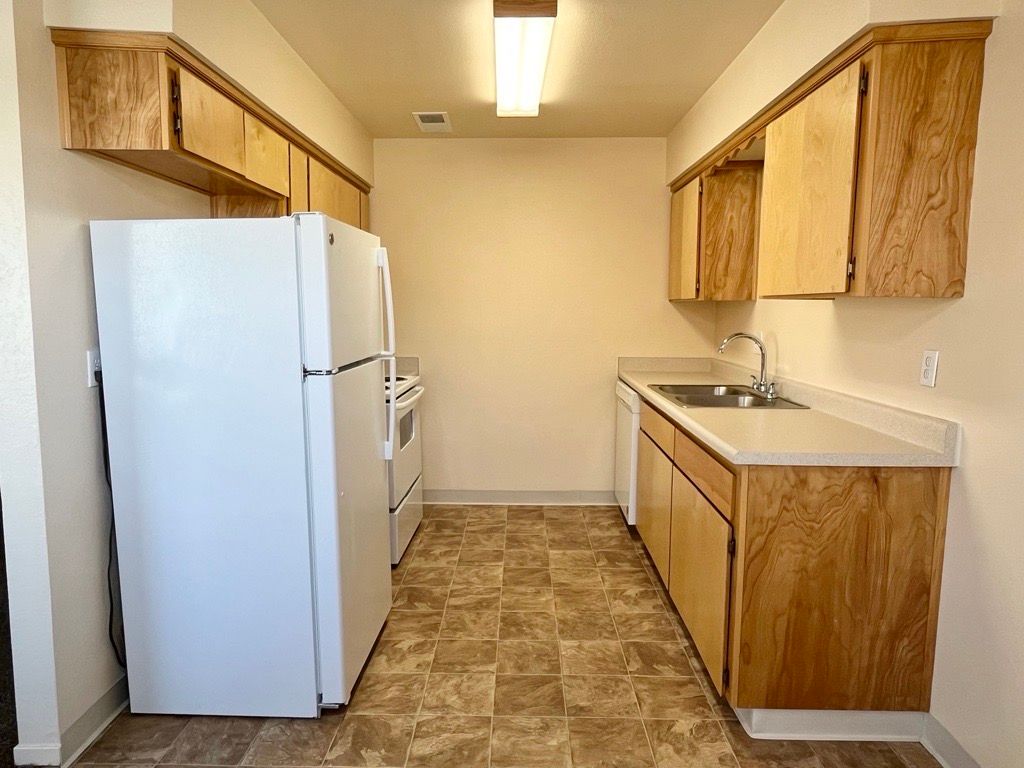 A galley kitchen with light wood cabinets, white appliances, and a neutral-toned floor.