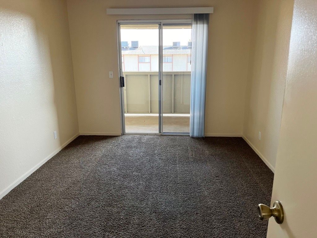 Empty room with brown carpet, sliding door to balcony, beige walls, and a partly visible door.