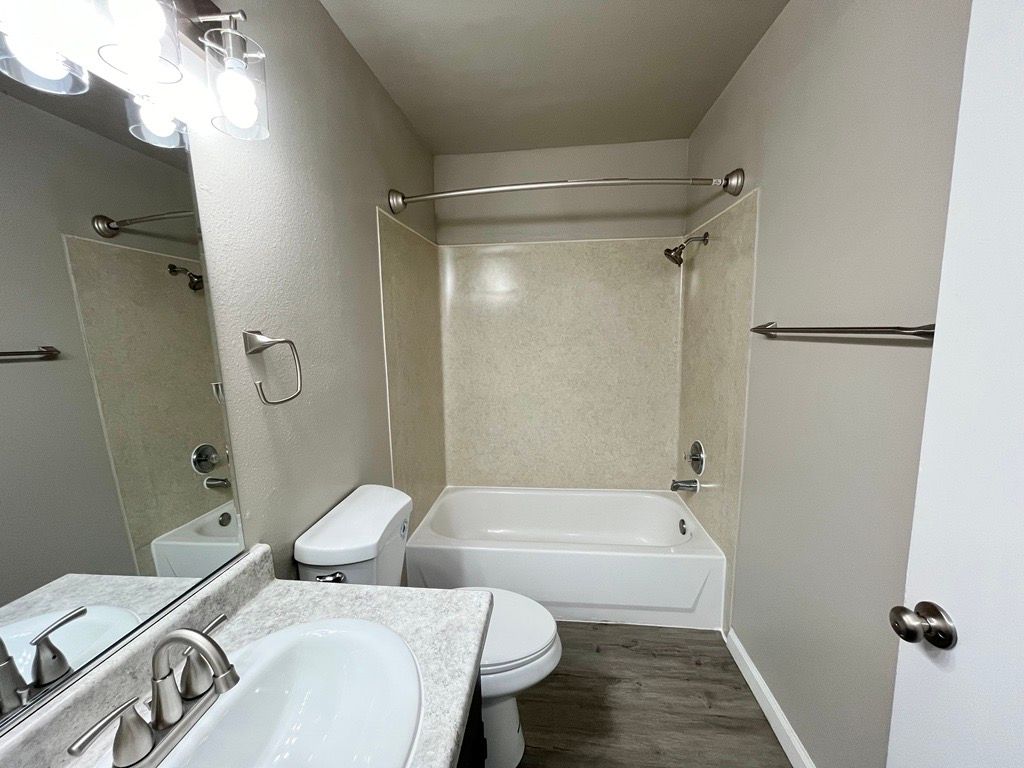 Bathroom with a white bathtub, toilet, and vanity. Grey walls, wood-look floor.
