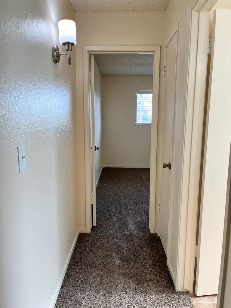Narrow hallway with beige walls, brown carpet, and doorway to a bedroom. A wall light is visible.