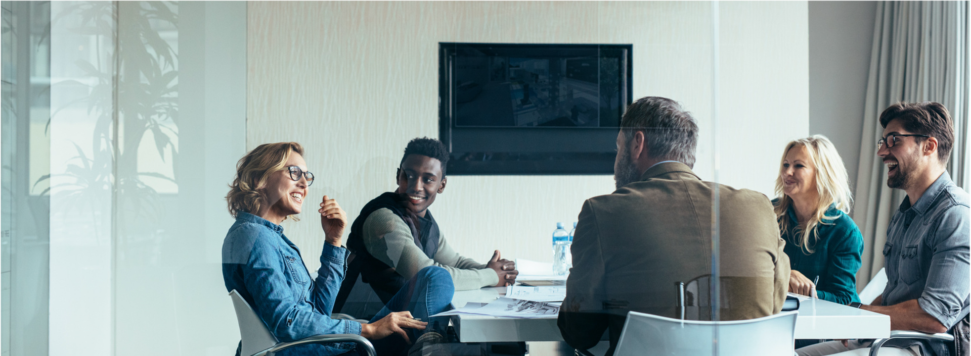 Meeting room with five people seated around a table, discussing in a bright office.