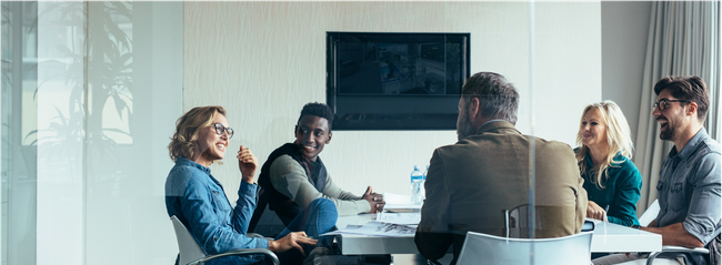 Meeting room with five people seated around a table, discussing in a bright office.