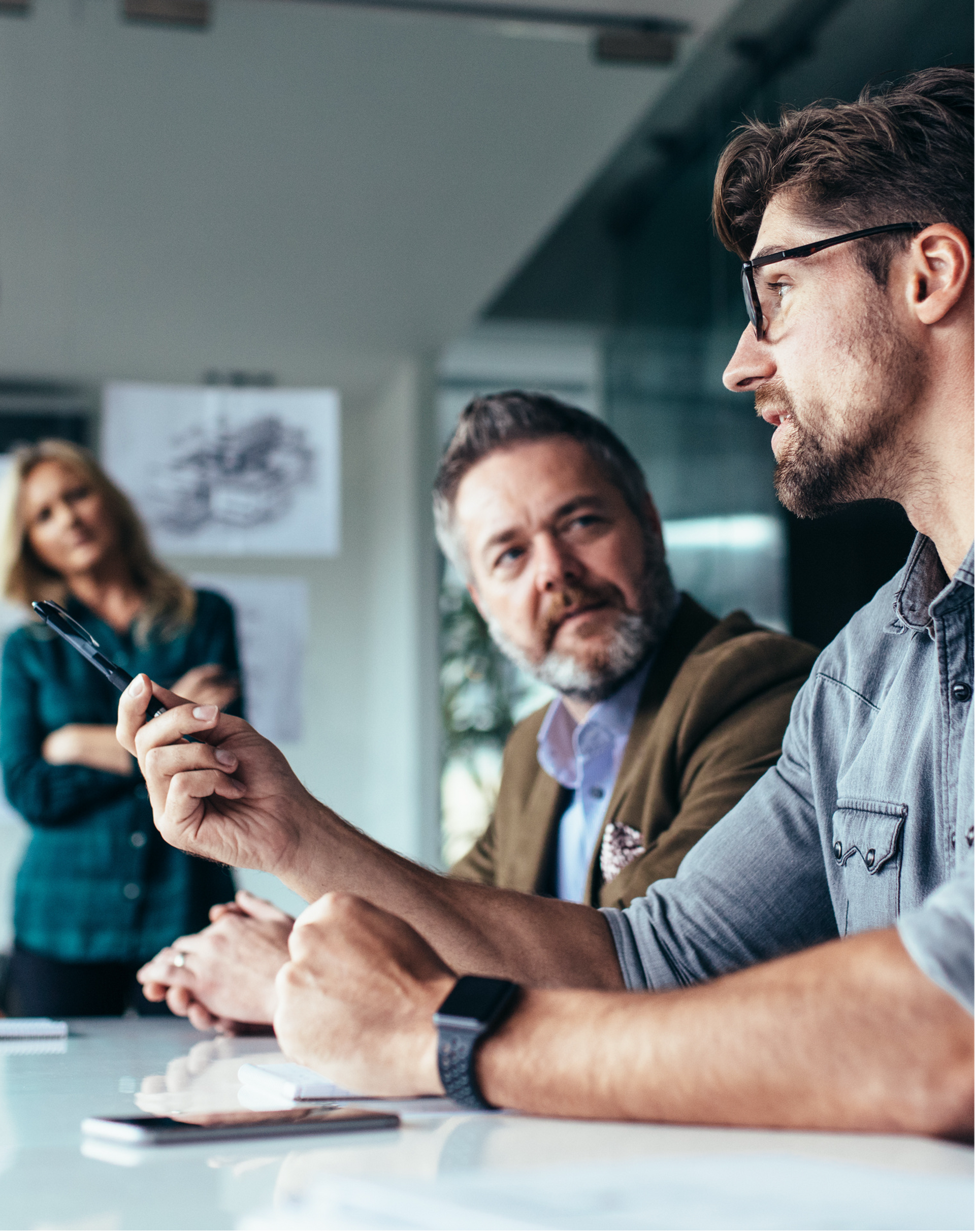 Three colleagues in a meeting, one gesturing while speaking at a table with notes.