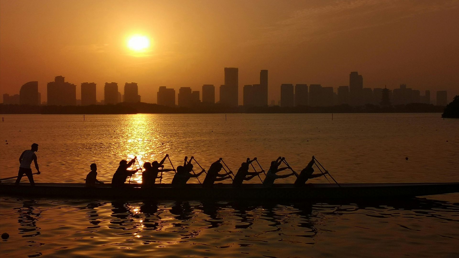 Rowers in silhouette on a sunset-lit river with a city skyline in the background
