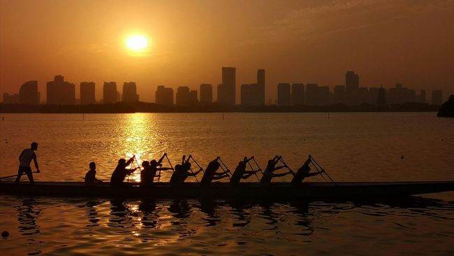 Rowers in silhouette on a sunset-lit river with a city skyline in the background