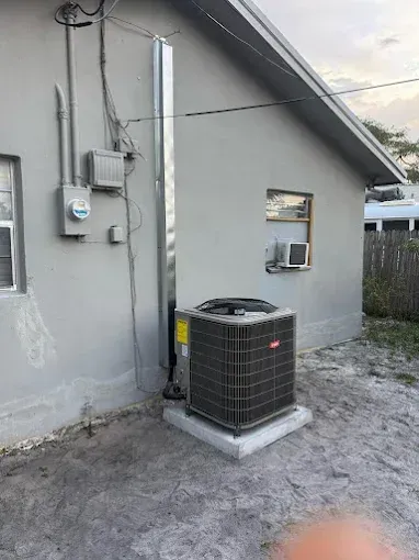 An AC unit sits outside a gray building with electrical components and a silver vent.
