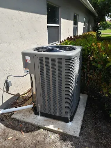 Gray air conditioning unit on a concrete pad next to a building and bushes.