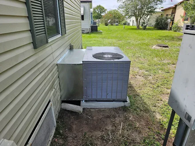 An air conditioning unit next to a house with beige siding and green shutters, on a concrete base.