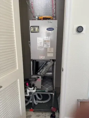 HVAC unit inside a closet, featuring a Carrier unit with pipes and a thermostat on the wall.