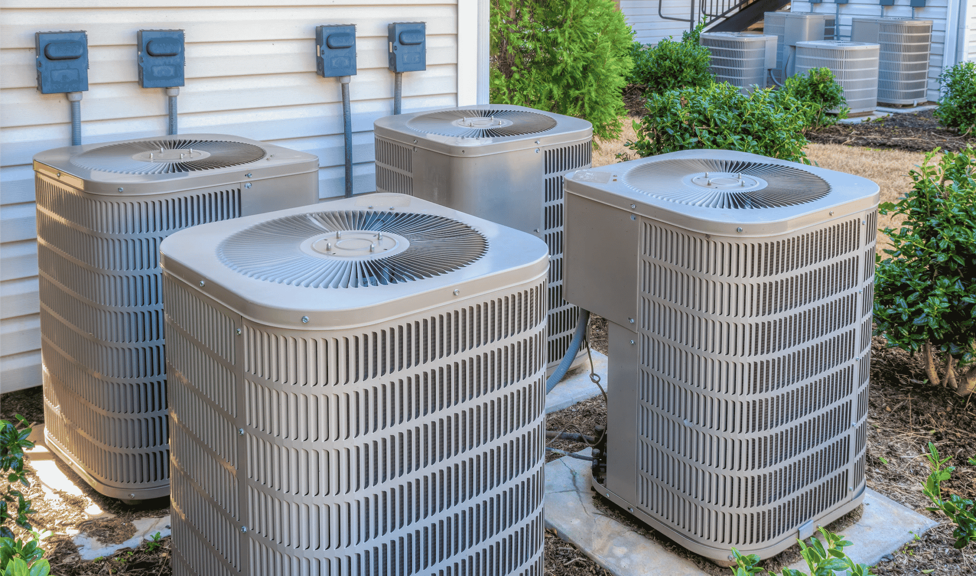 Four outdoor air conditioning units near a building, with electrical boxes visible.