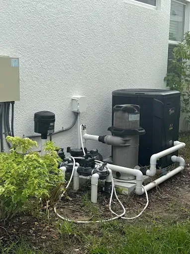 Pool equipment against a white stucco wall: pump, filter, heater, and valves with white piping and electrical box.