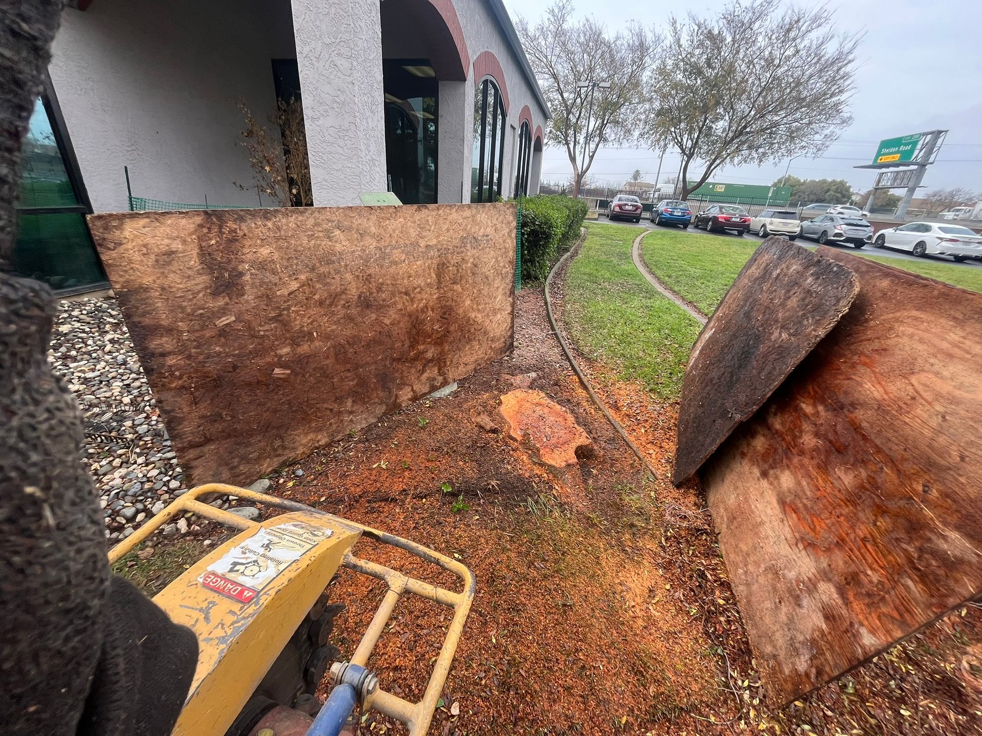 A person operating a yellow tamper in front of a building with a damaged wall and landscaping.