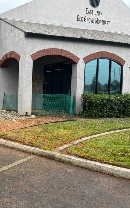 East Lawn Elk Grove Mortuary building with arched entryway and window, green foliage, cloudy sky.