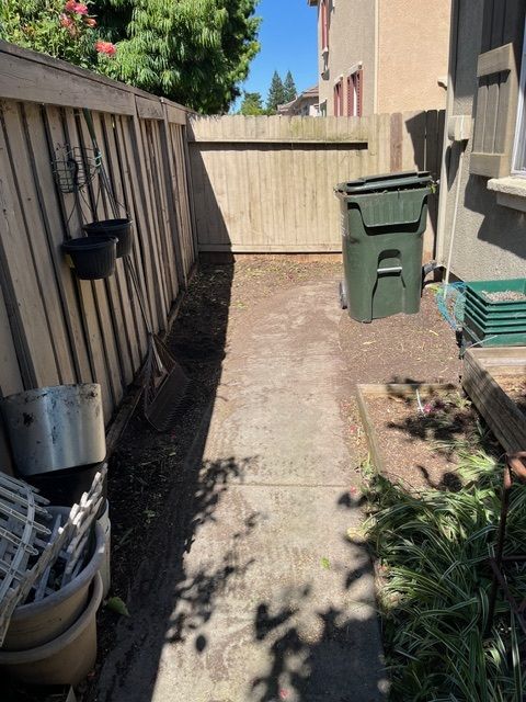 Narrow concrete path between wooden fences, trash can, and house with plants. Narrow concrete path between wooden fences, trash can, and house with plants.