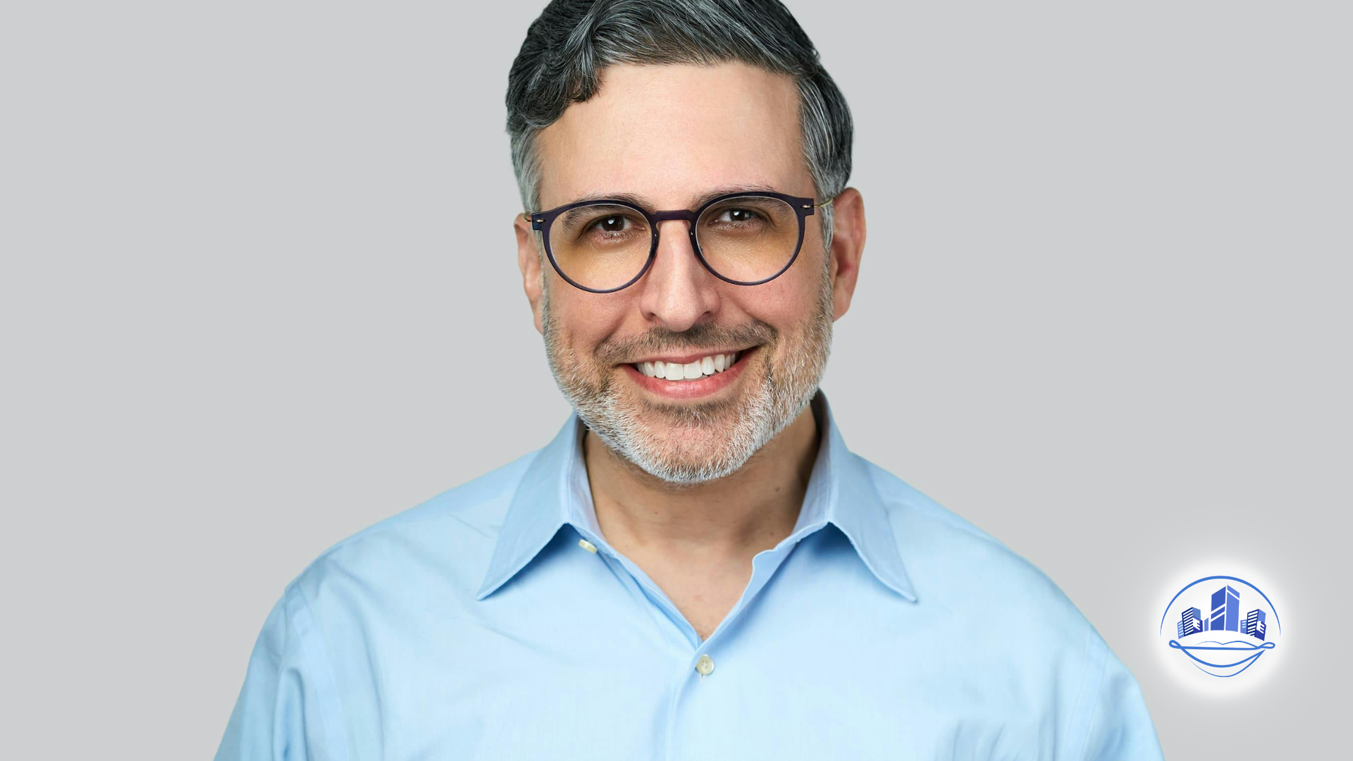 Man with glasses smiling, wearing a blue shirt against a gray background.
