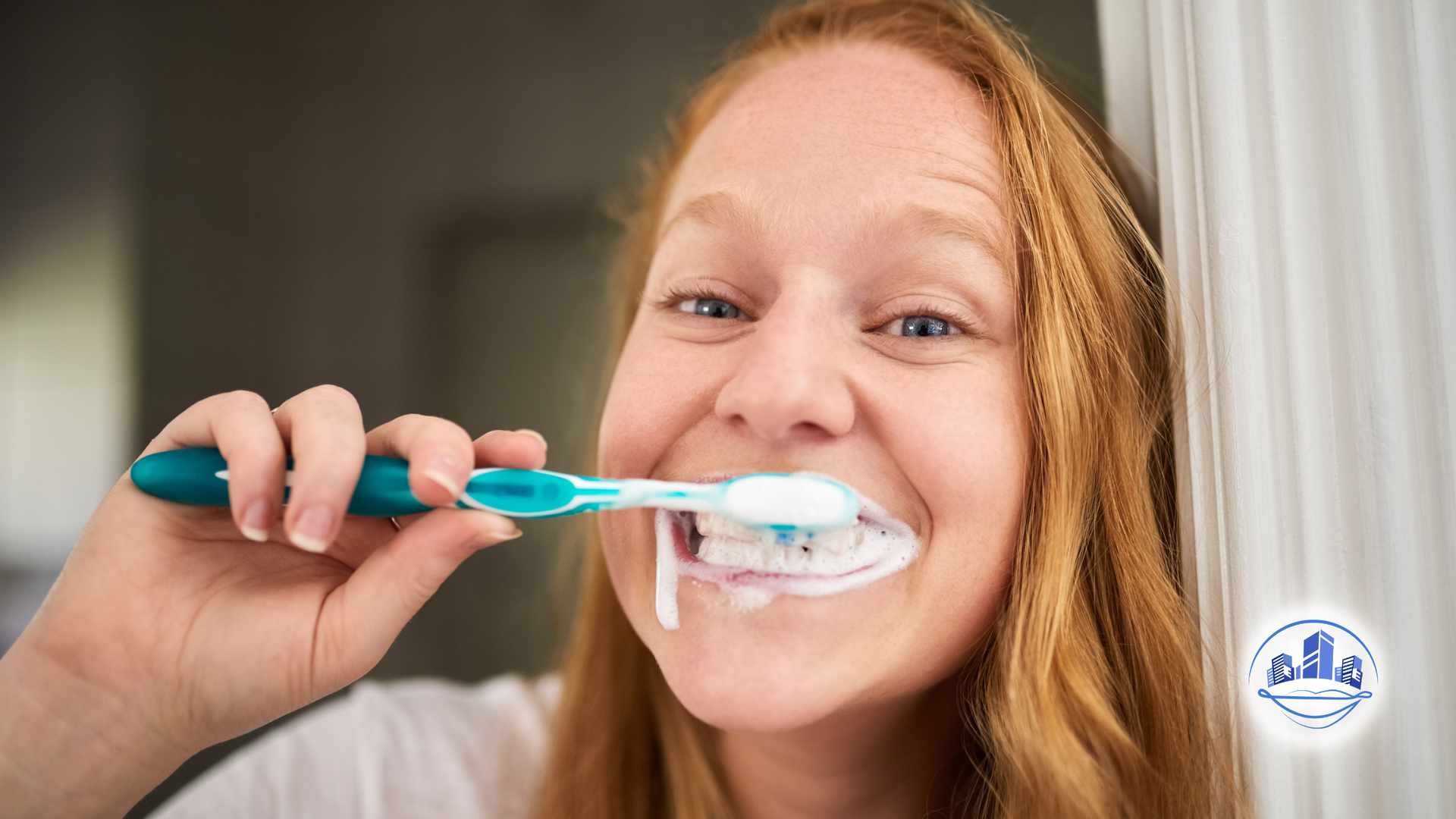 Woman with red hair brushes teeth with toothpaste in a bathroom, smiling.