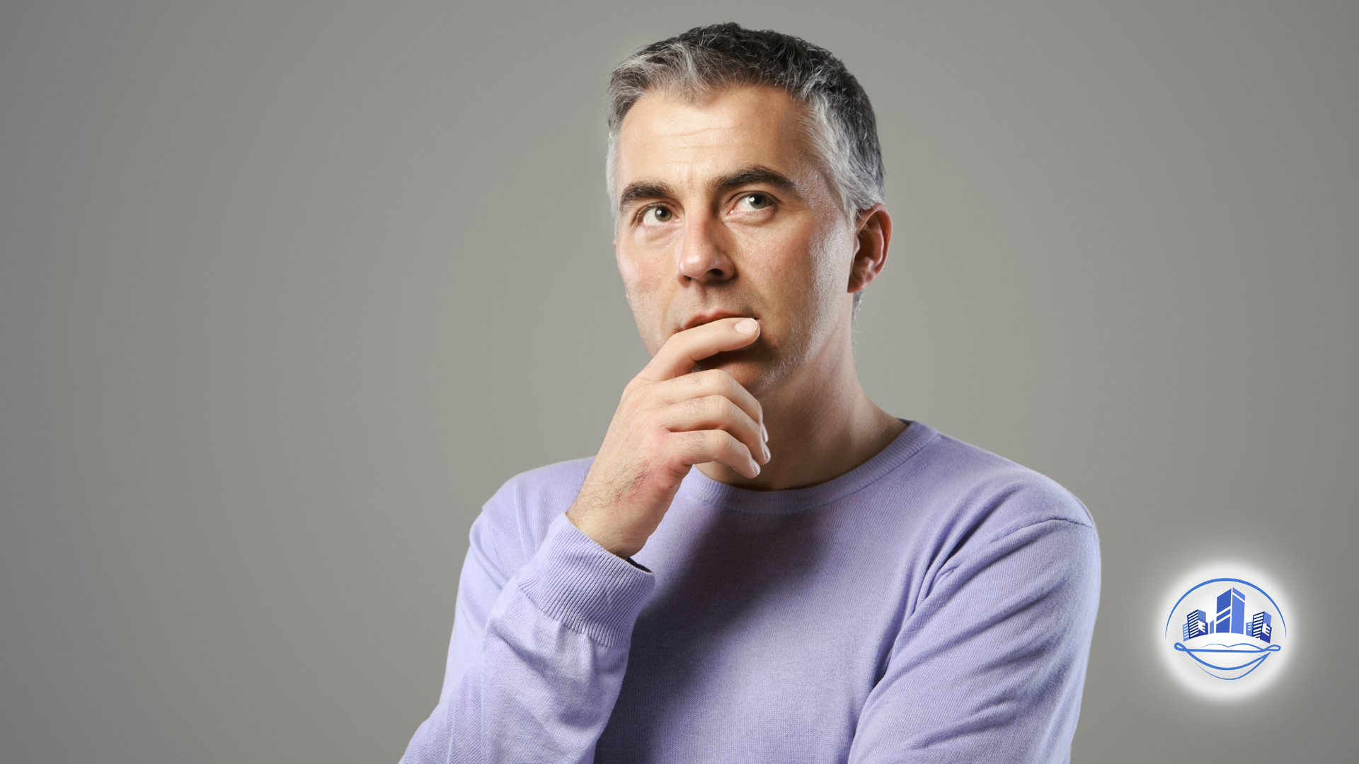 Man in light purple sweater, hand on chin, thinking, against a gray background.