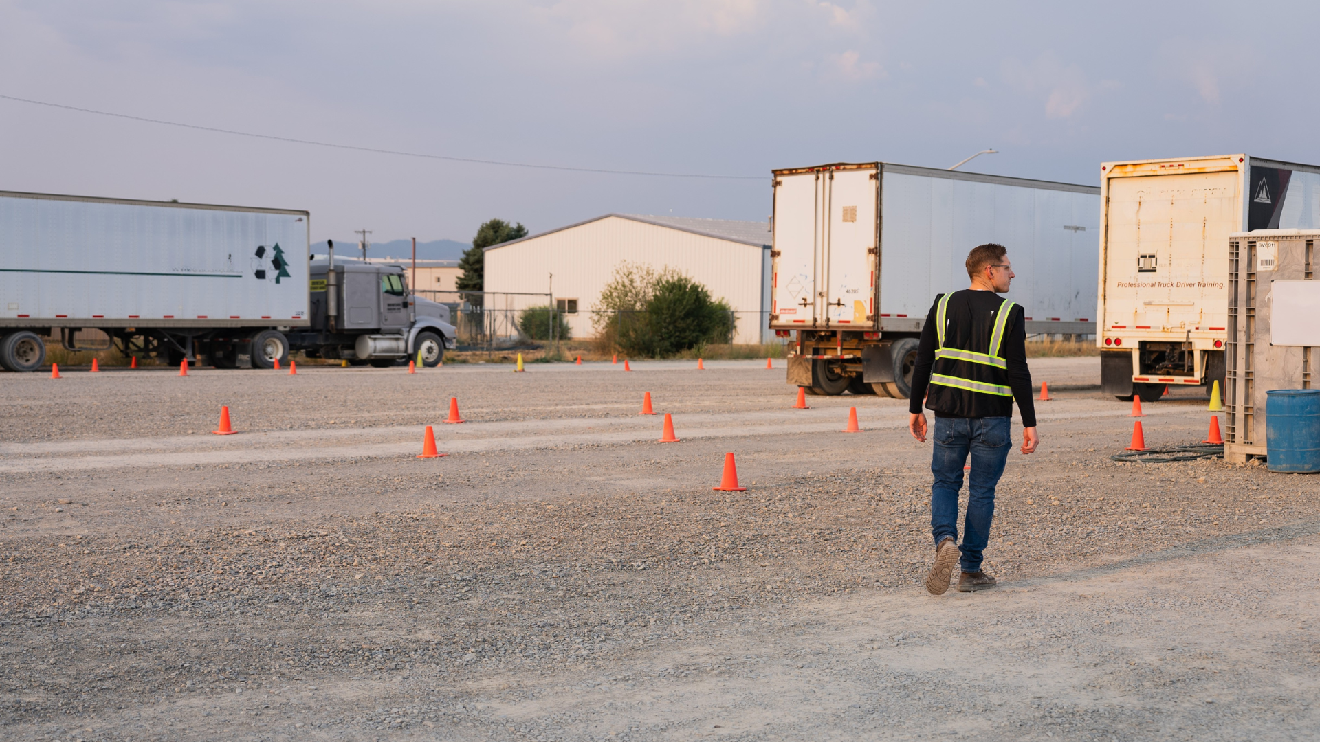 Man in safety vest walks past parked semi-trucks and cones in gravel lot doing CDL training