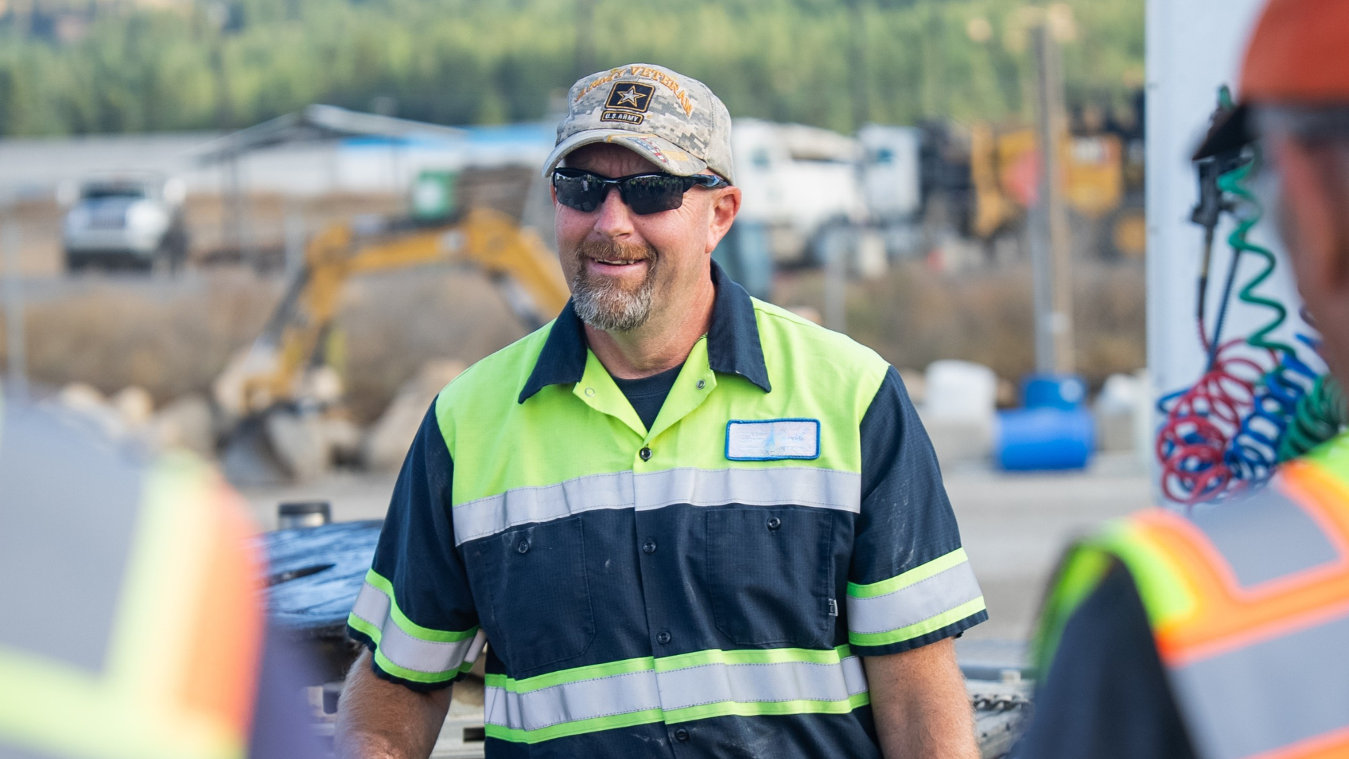 Man in safety vest smiling at a worksite, wearing sunglasses and a US Army hat