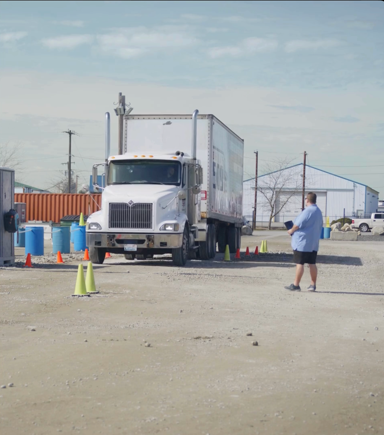 Truck driving training: white semi-truck driving through cones. Instructor watches in a dirt lot on a sunny day. CDL training with state examiner eldt course