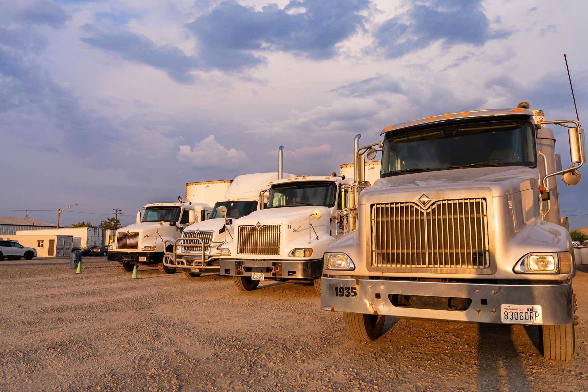 A row of semi trucks are parked in a CDL training gravel lot