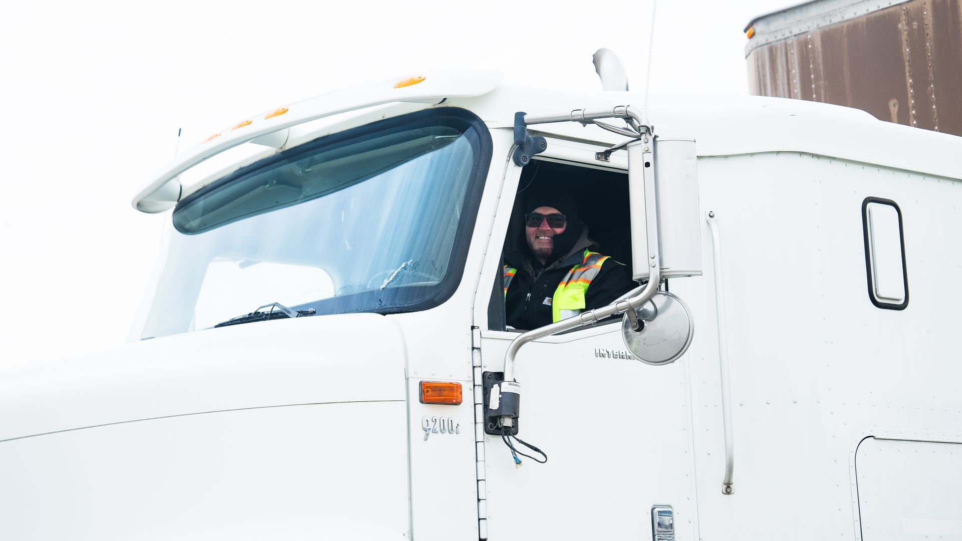 Truck driver in a white semi-truck wearing a black hat and yellow vest, smiling in a snowy environment.