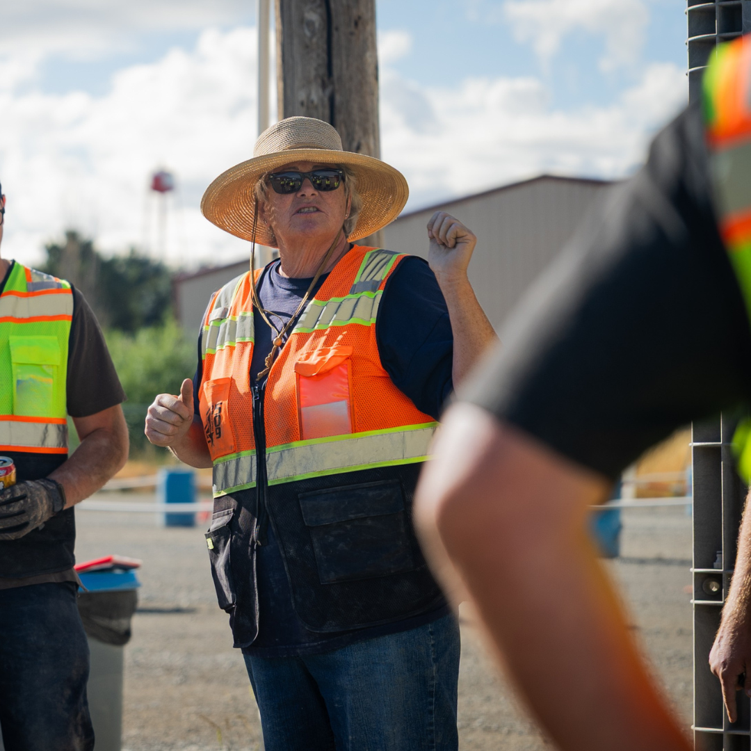 Woman in safety vest and straw hat gestures while talking with construction crew outdoors.