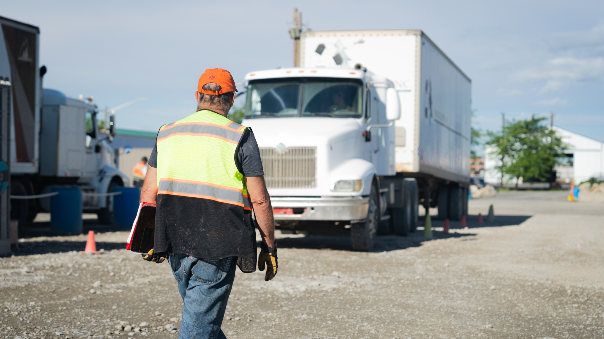 Worker in safety vest walking towards white truck, carrying a clipboard, outdoors.