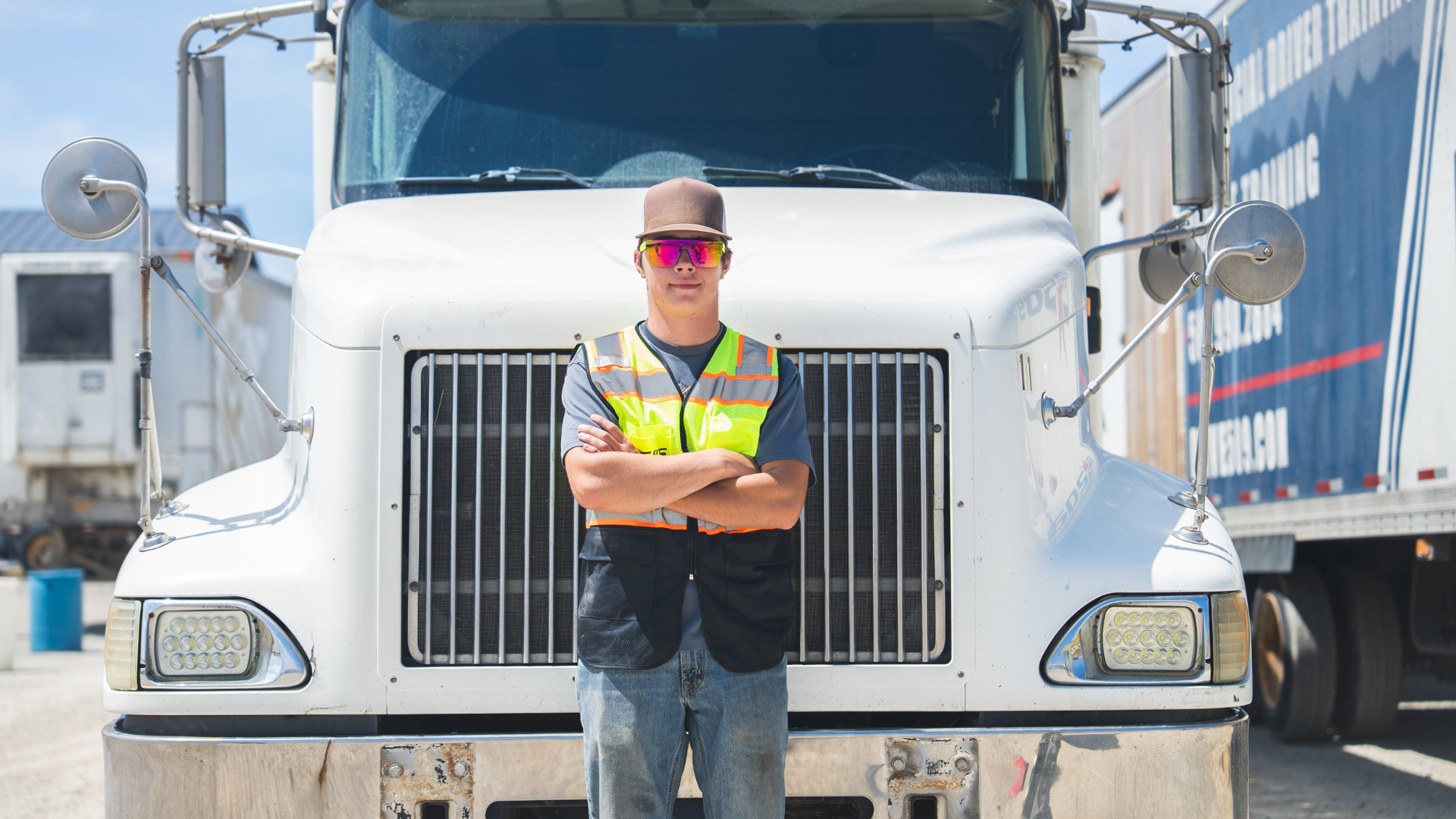 Truck driver in vest and hat stands with arms crossed in front of a white semi-truck.