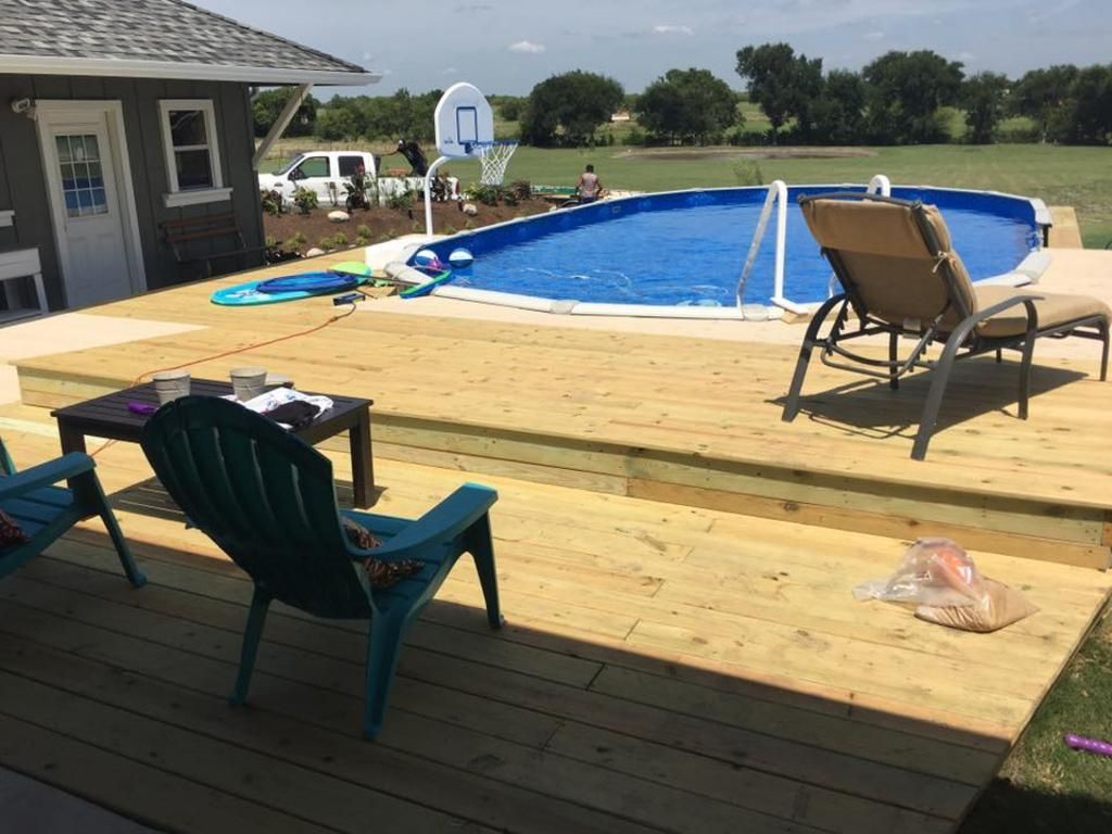A large swimming pool is surrounded by a wooden deck with chairs and a table.