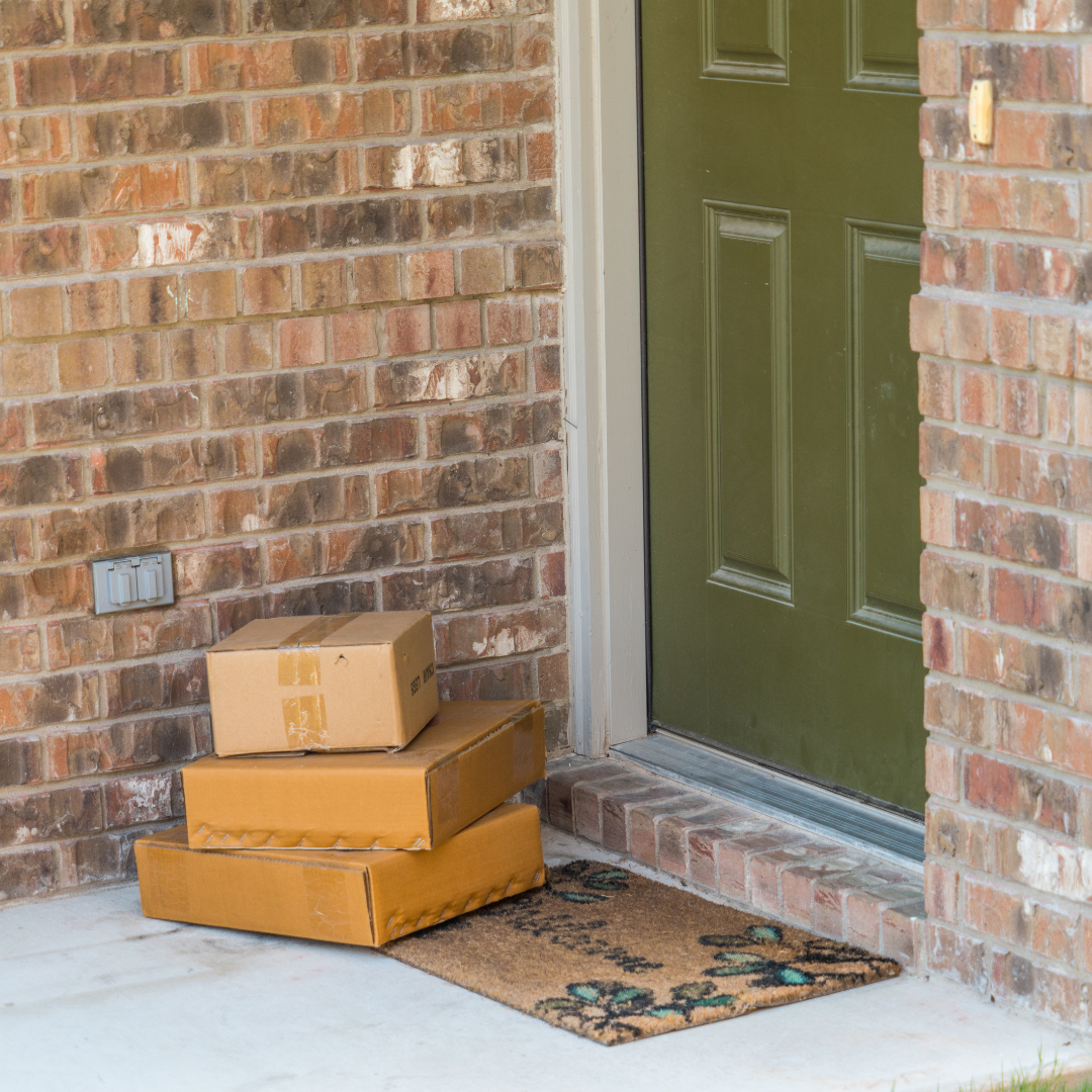 Three cardboard packages sit outside a green door on a porch with a brick wall.