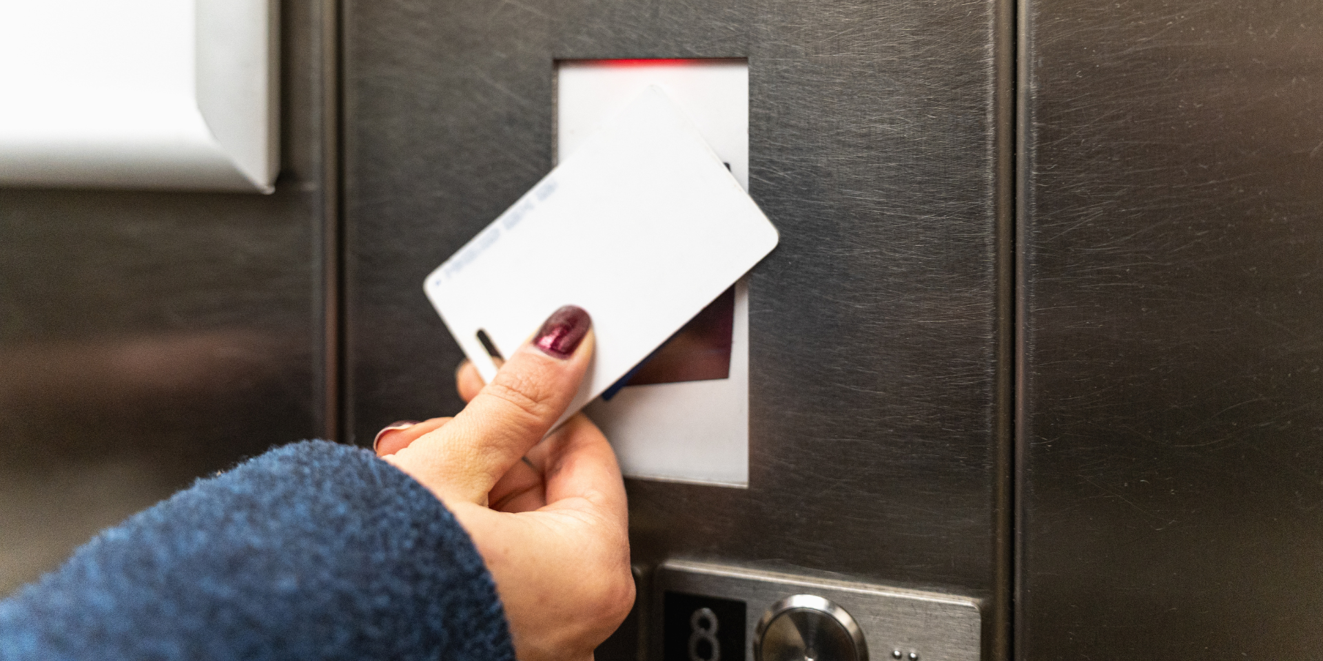 A hand holds a white plastic access card up to a card reader panel inside an elevator.