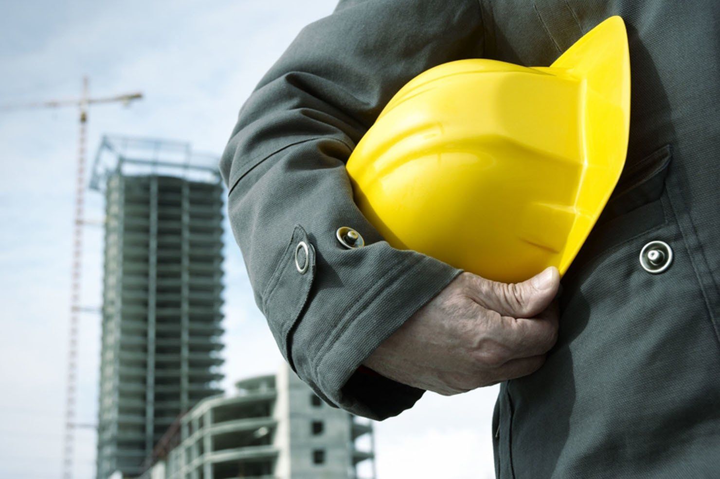 Construction worker holding yellow hard hat, building under construction in background.