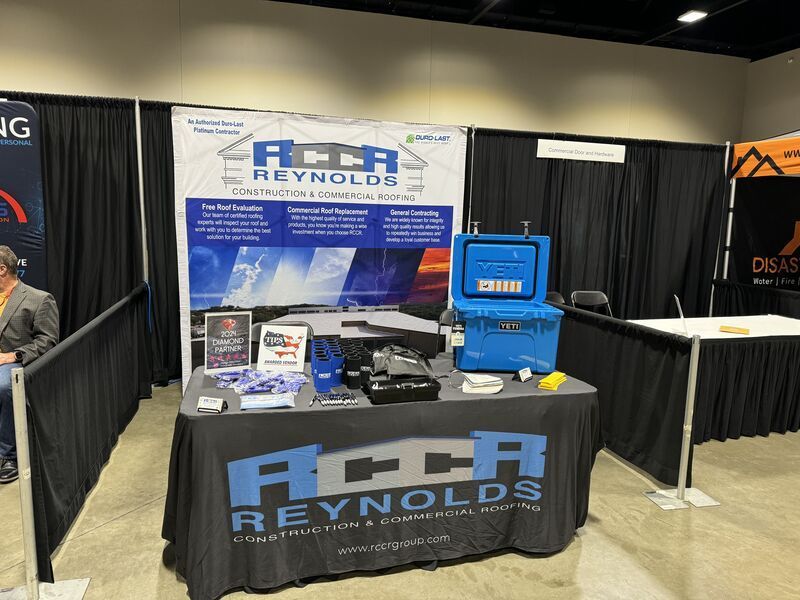 A trade show booth for Reynolds Construction & Commercial Roofing featuring a table with a blue cooler and company banner.