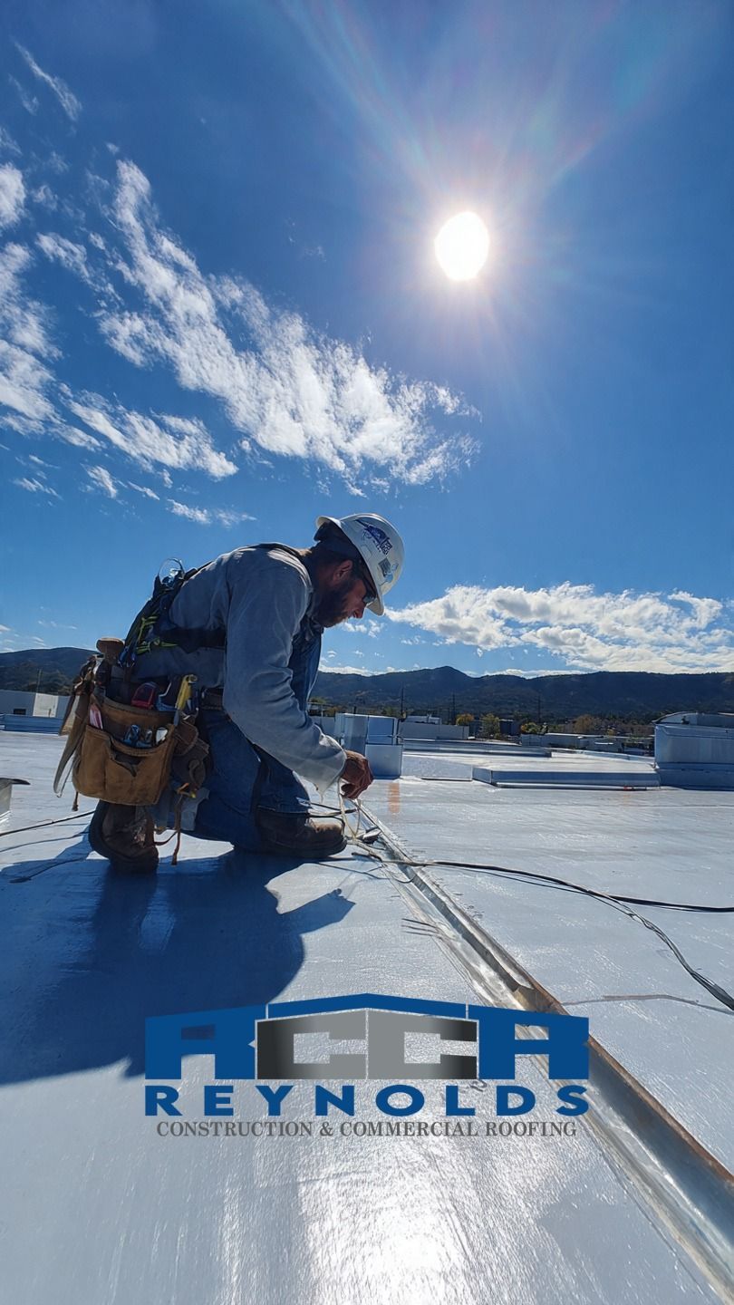 A worker in a hard hat and tool belt kneels on a white roof, applying a seam seal, under a bright sun and blue sky.