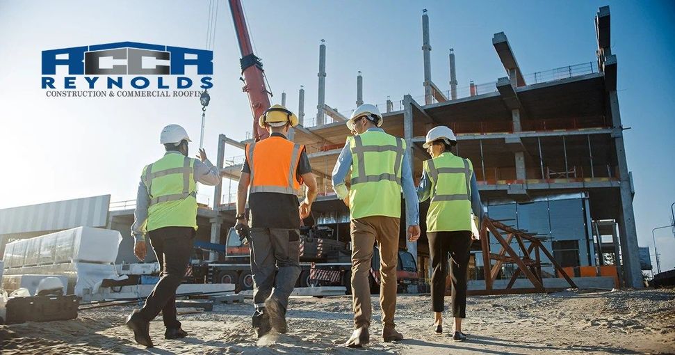 Four construction workers in safety vests and hard hats walk across a sunlit construction site toward an unfinished frame.