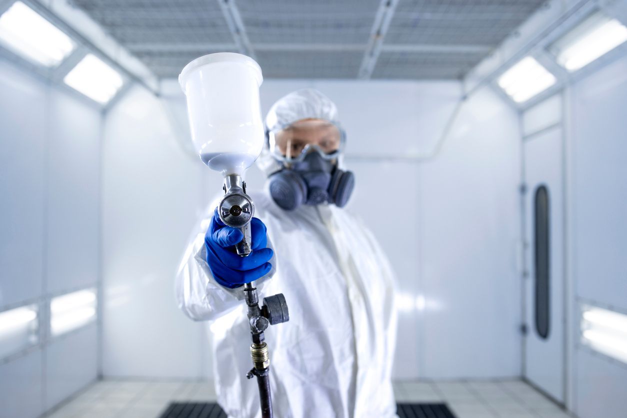 A person in protective gear and a respirator holds a paint spray gun toward the camera in a white professional booth.