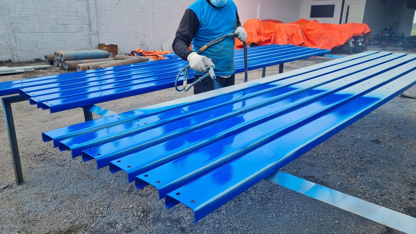 A person in a blue shirt spray-paints rows of long, bright blue metal beams outdoors.
