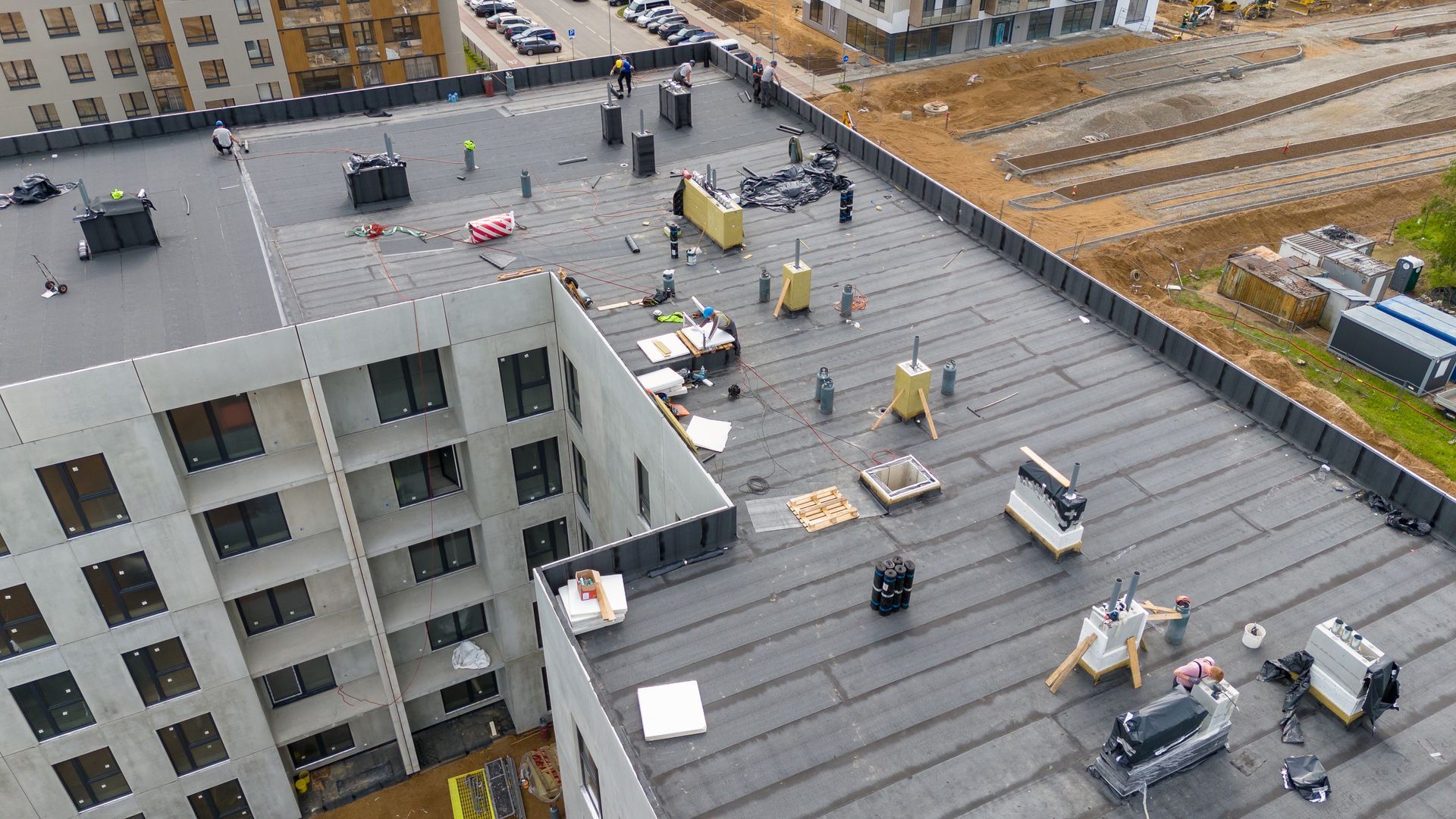 Aerial view of construction workers installing a black membrane roof on a multi-story concrete apartment building.