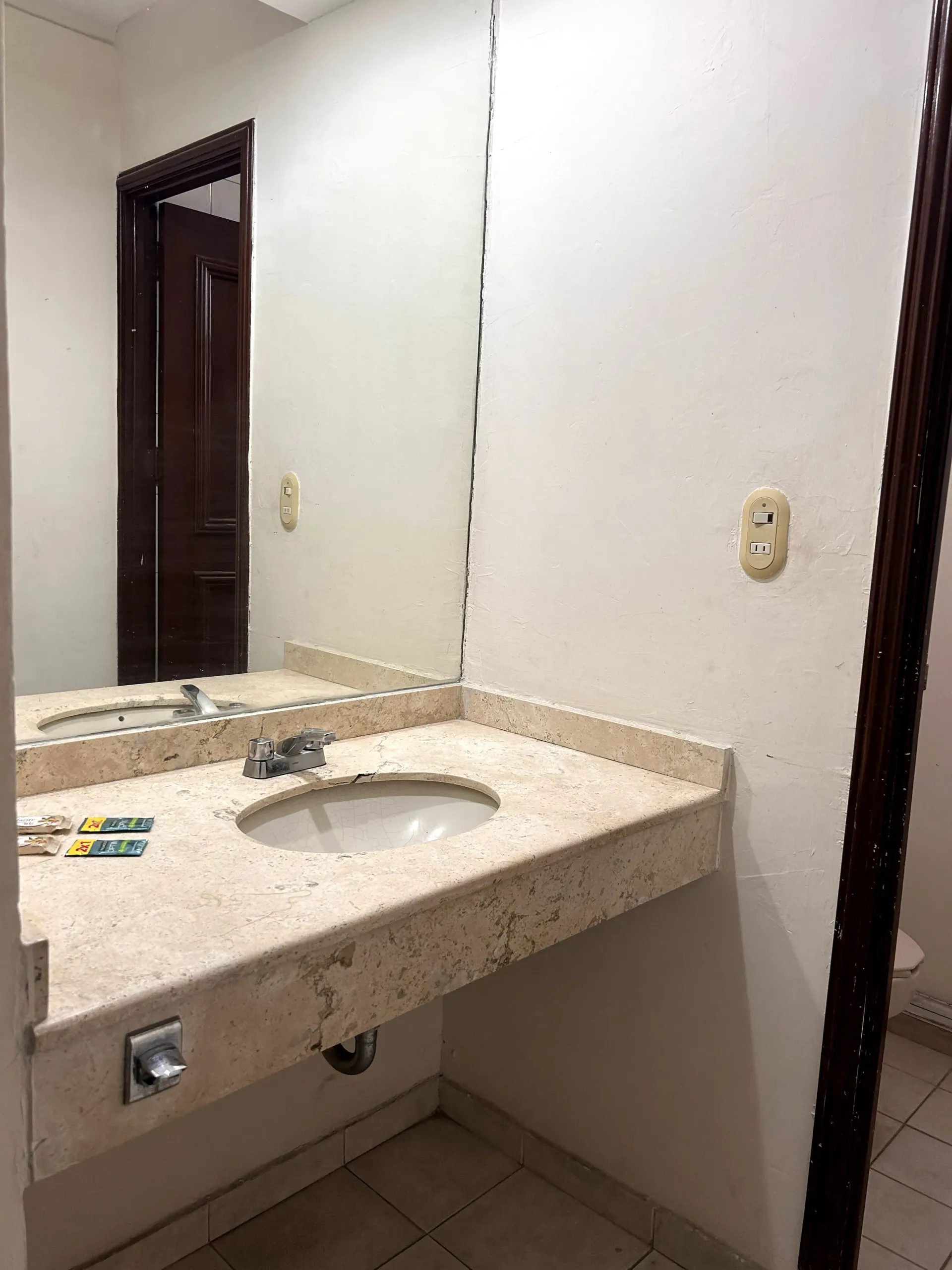 Un tocador de baño de mármol beige con lavabo, espejo y toma de corriente, sobre paredes lisas de color claro.