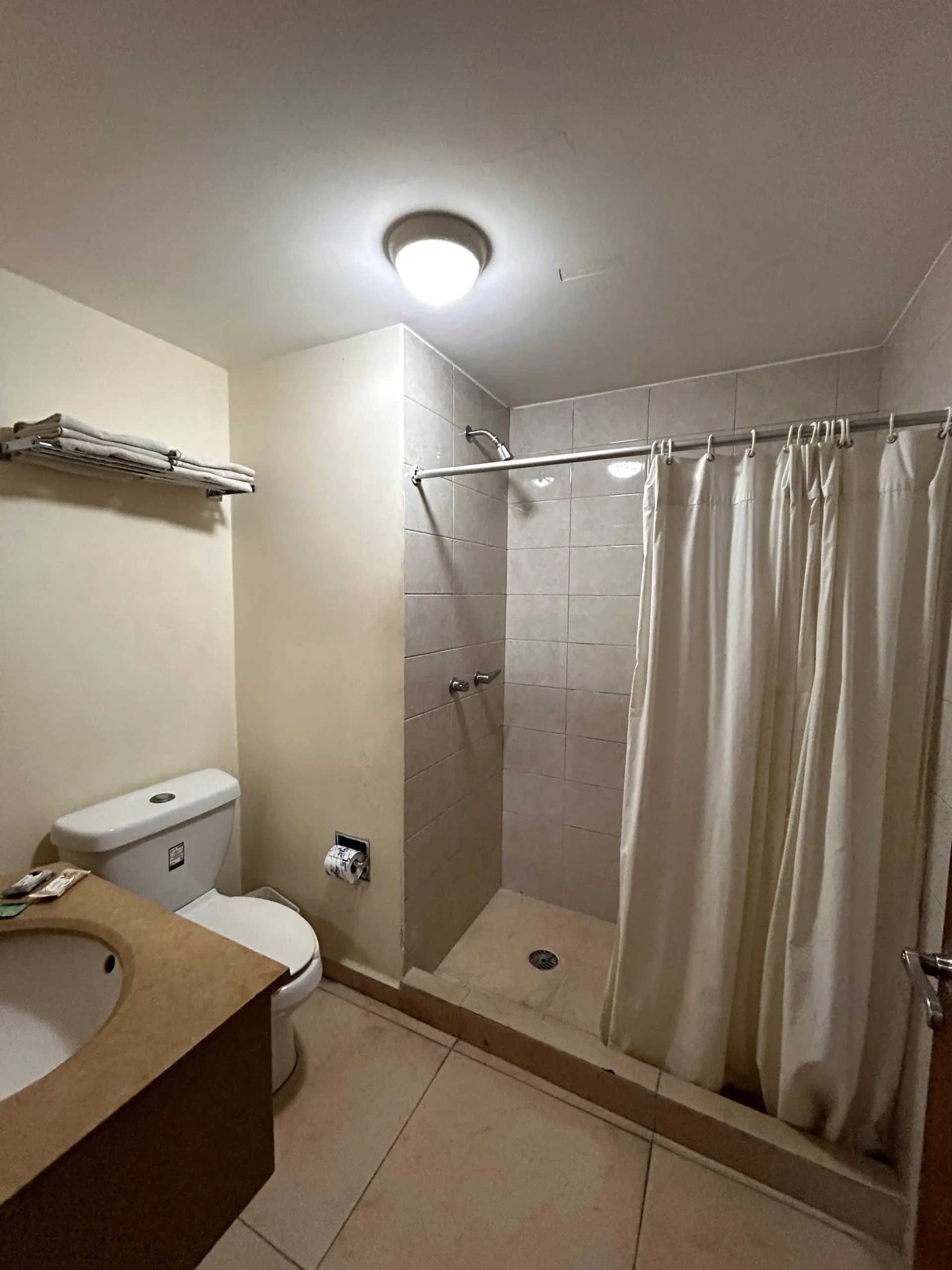 Un baño con tocador de color beige, inodoro y ducha a ras de suelo con una cortina beige detrás de una repisa elevada.