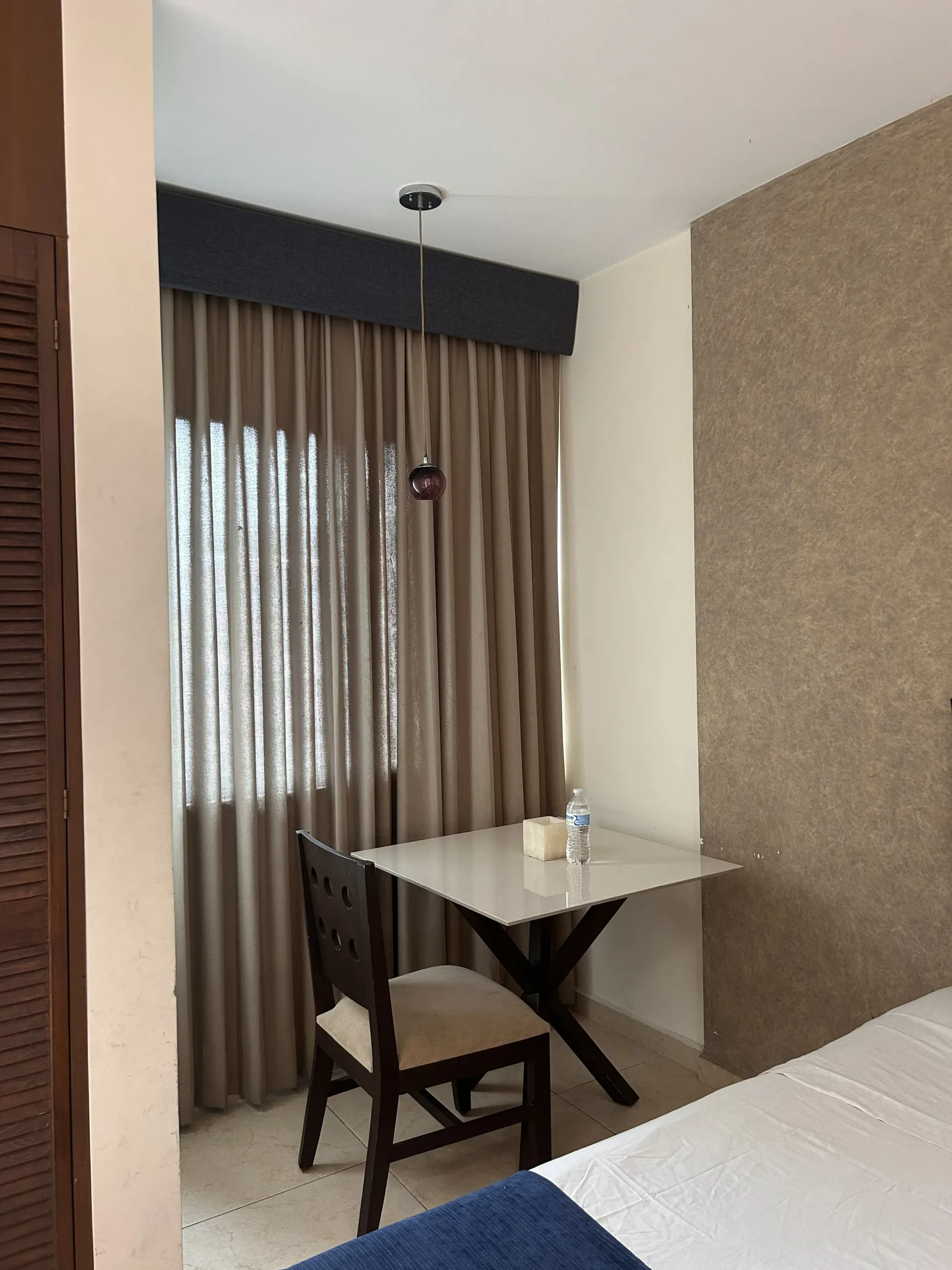 Una mesa cuadrada con una silla cerca de una ventana con cortinas beige hasta el suelo y una cenefa gris oscuro.