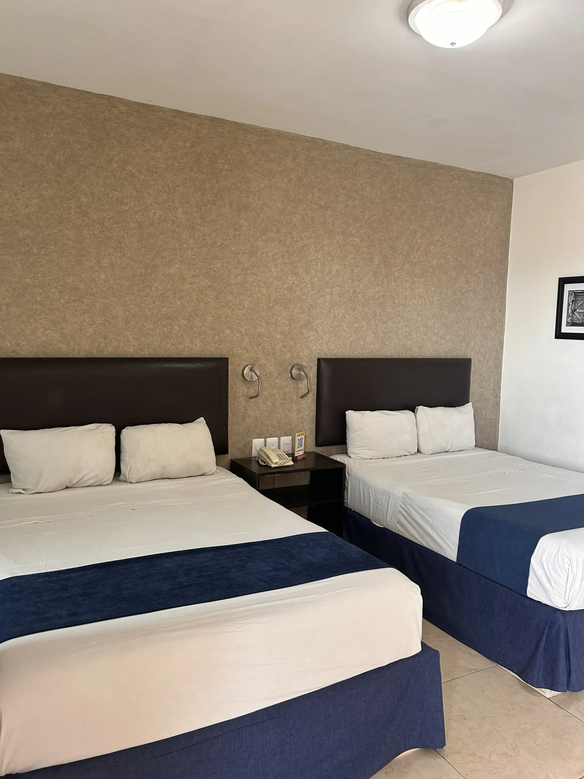 A hotel room with two queen beds, dark headboards, white linens, blue bed runners, and a textured beige accent wall.
