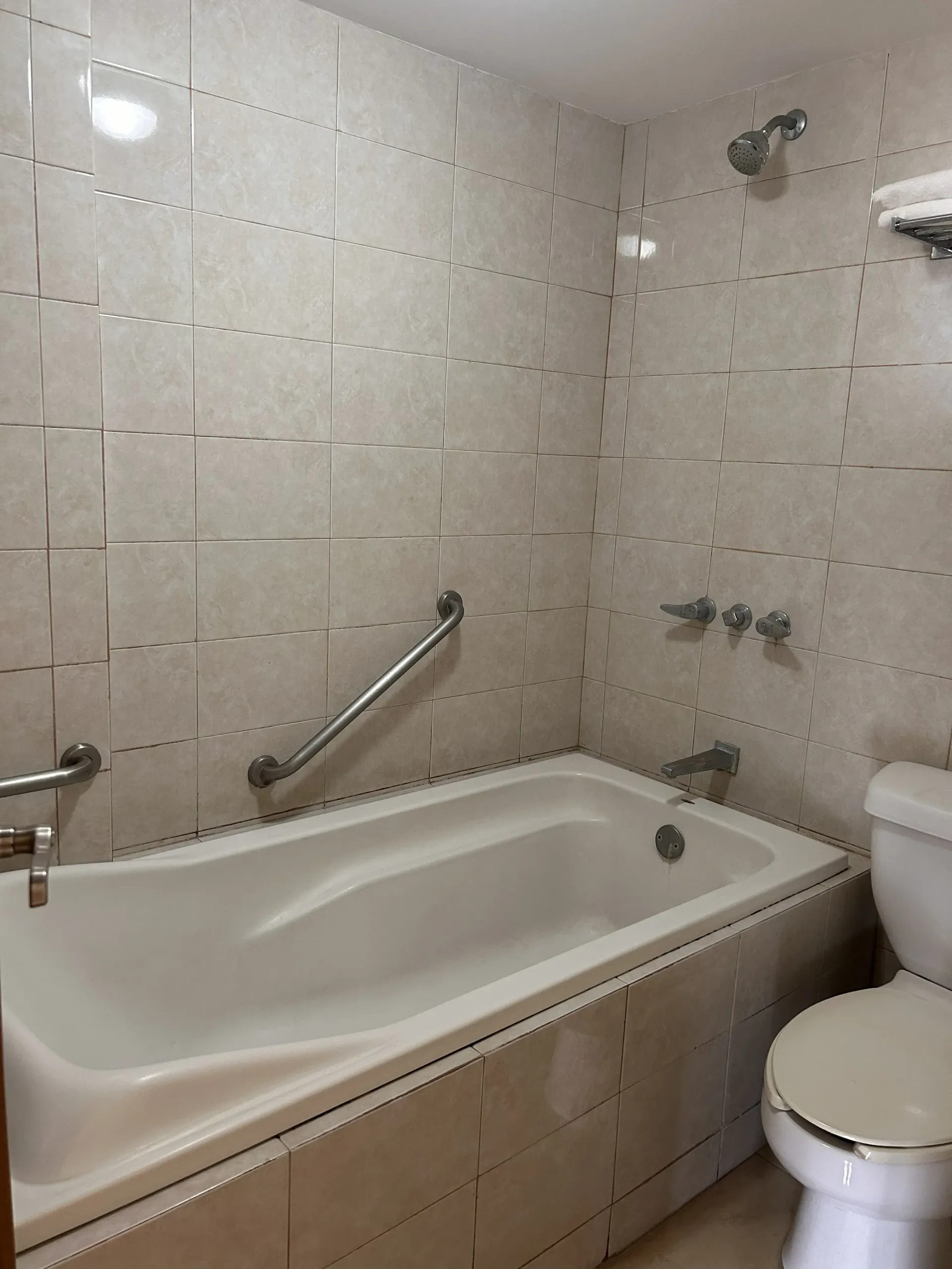 Un baño con azulejos de color beige, bañera blanca con barra de apoyo metálica y un inodoro estándar.