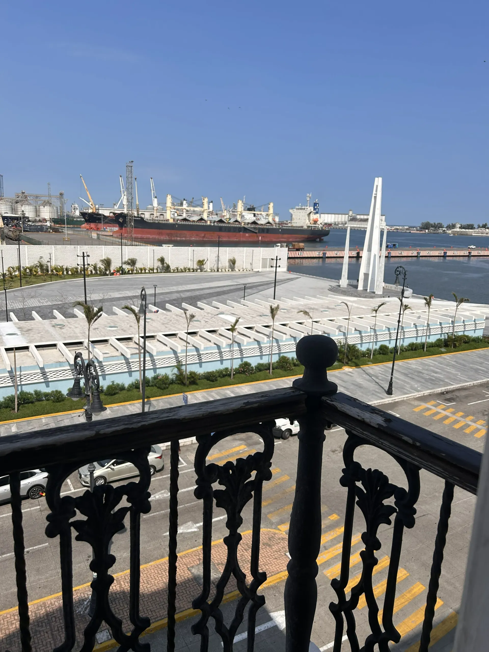 A balcony railing overlooks a harbor with a large cargo ship, a paved plaza, and a tall white monument under a blue sky.