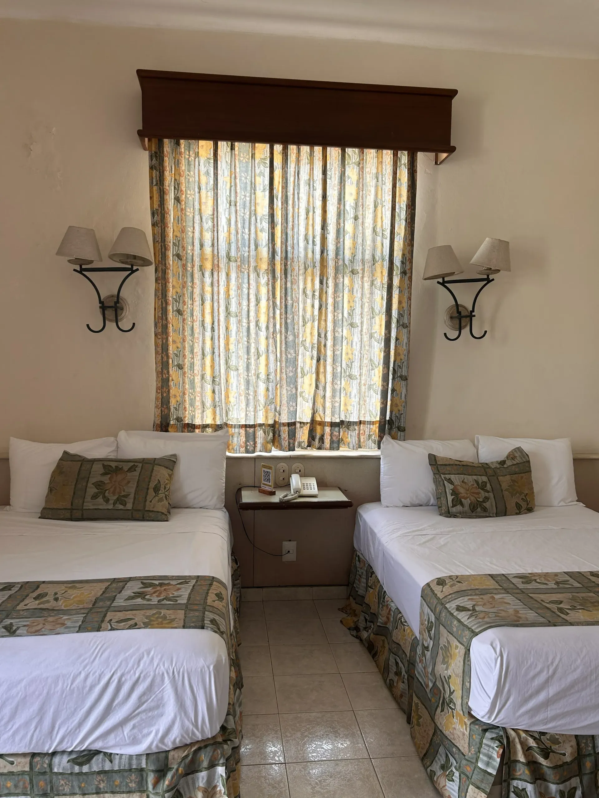 Dos camas con faldones y cabeceros estampados en una habitación con una ventana central, cortinas beige y apliques de pared.