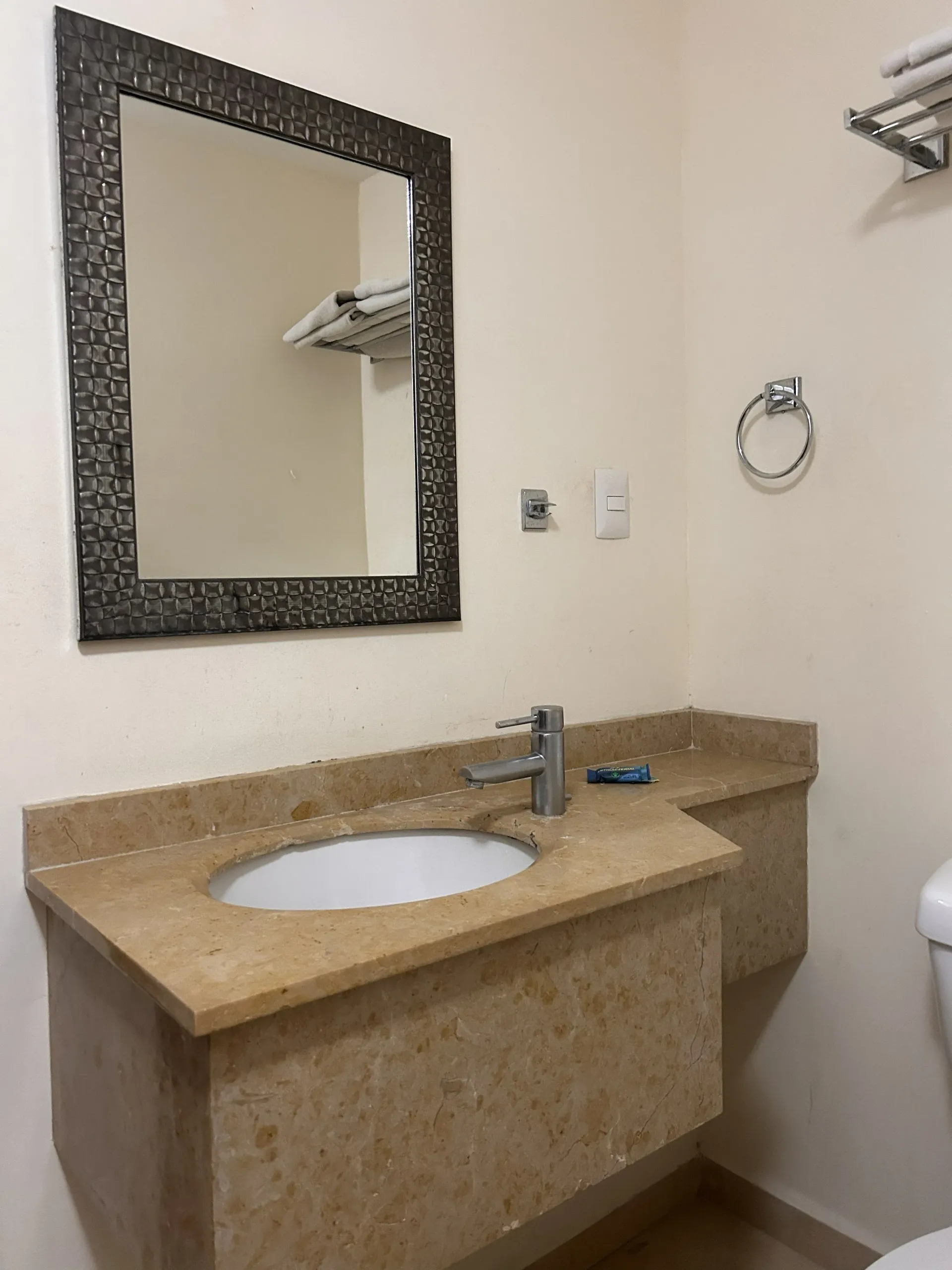 Un mueble de baño con encimera de piedra beige, lavabo blanco, grifo plateado y espejo rectangular con marco oscuro.