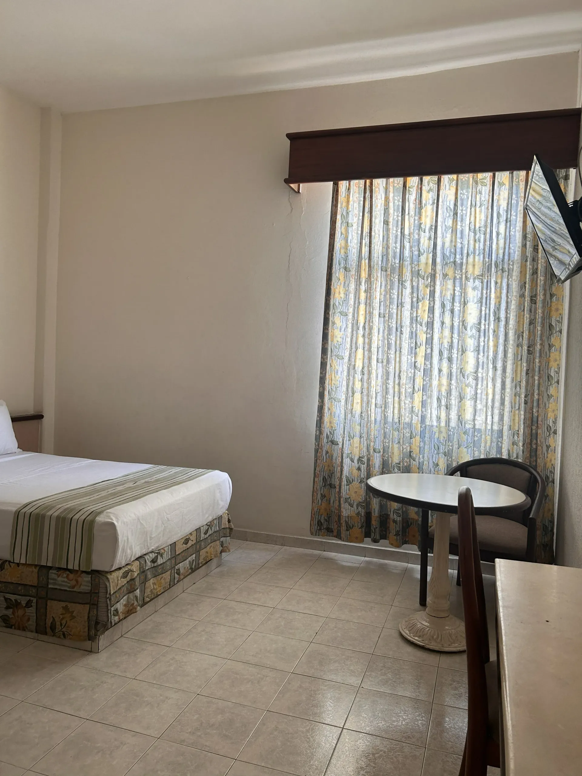 Una habitación de hotel con una cama individual, una pequeña mesa redonda con una silla y cortinas estampadas en la ventana.