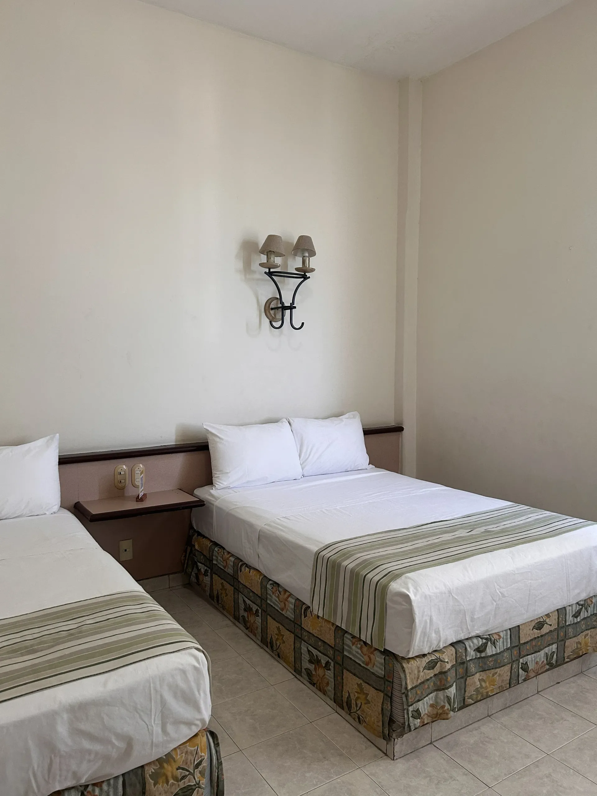 Dos camas blancas con caminos de mesa estampados en una habitación sencilla, con una lámpara.